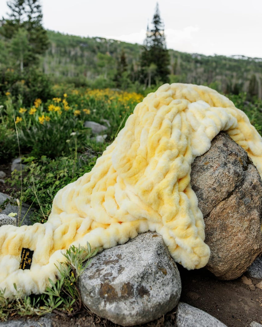 A stretchy blanket in pale yellow and cream is draped over large rocks in an outdoor mountain setting with wildflowers and trees in the background.