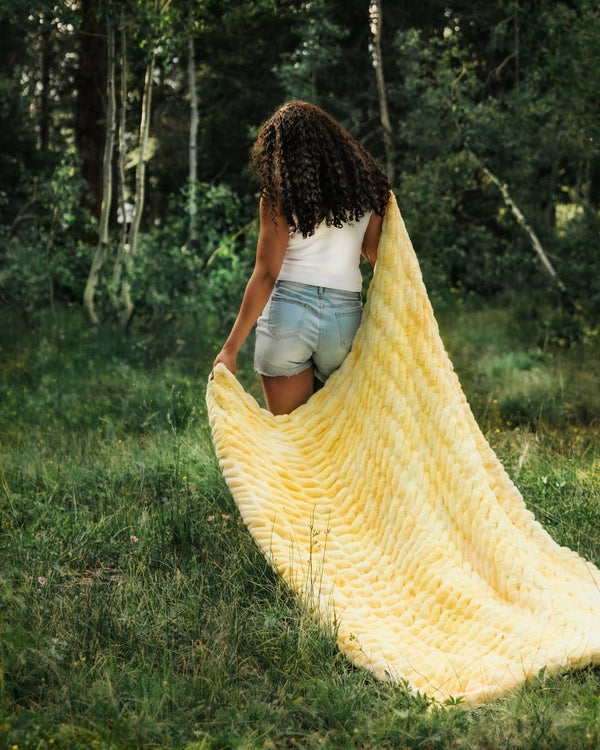 A woman stands in a grassy forest clearing holding up a long pale yellow blanket, showing its thick stretchy fabric.