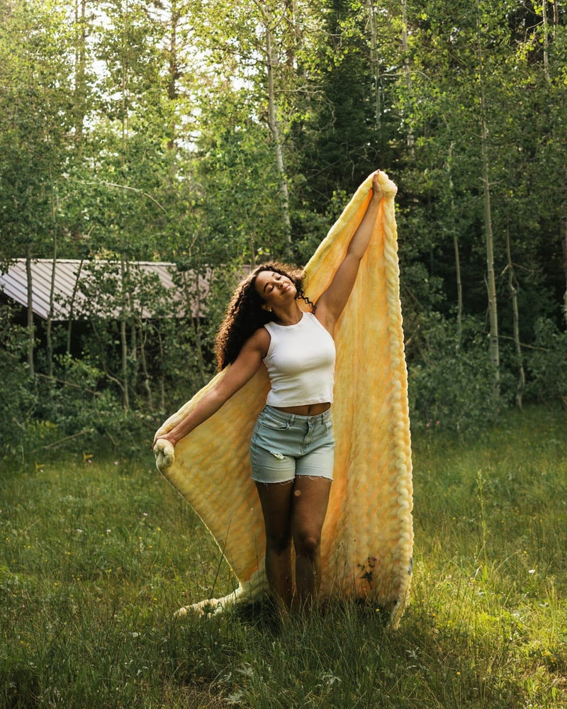A woman standing in a grassy outdoor area surrounded by trees, holding up a large yellow blanket or throw with both arms.