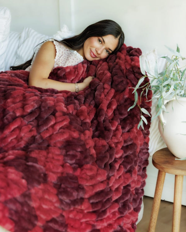 A woman lies on a bed covered with a chunky, Hugs Diamond Red Blanket  in deep red and burgundy tones. The room has white bedding, a small wooden side table with a white vase of greenery, and a soft neutral wall background.