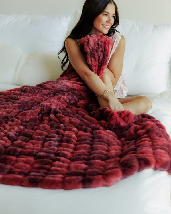 A woman sitting on a white bed wrapped in a chunky, Hugs Diamond Red Blanket. The blanket features a gradient of deep red and burgundy tones.