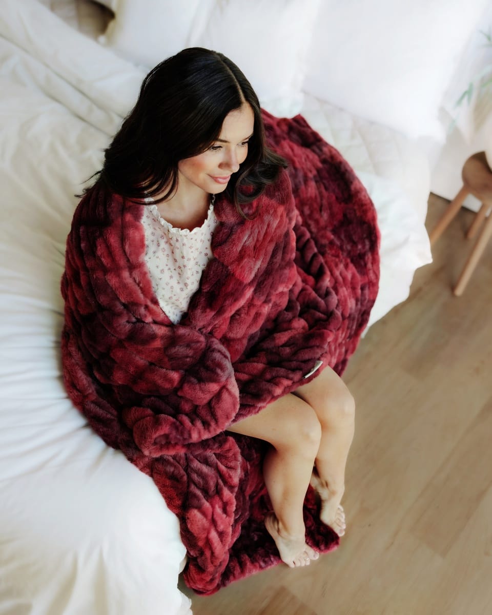 A woman sits on a white bed wrapped in a chunky, Hugs Diamond Red Blanket. The blanket has a rich burgundy color with subtle tonal variations from the texture and lighting.