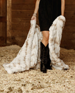 A person standing in a barn-like setting with wood walls and straw floor, holding up a large white and beige fur throw or blanket. 
