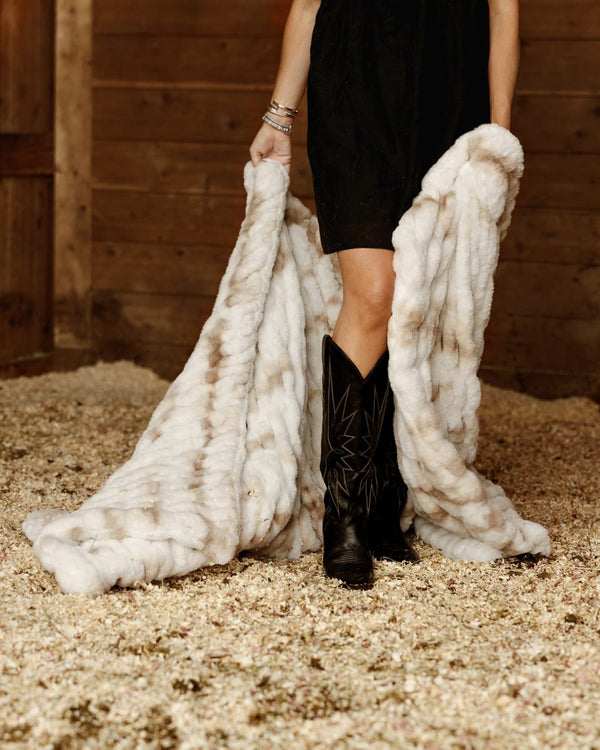 A person standing in a barn-like setting with wood walls and straw floor, holding up a large white and beige fur throw or blanket. 