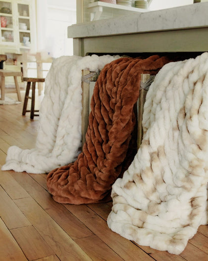 Three fluffy throw blankets in white, rust brown, and cream are draped over the chairs by a kitchen island in a bright, modern kitchen.