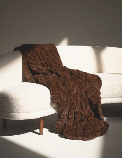 Hugs Hot Fudge Blanket draped elegantly over a stylish cream sofa, showcasing its rich brown color and plush texture.