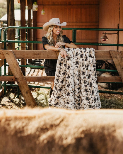 A woman in a cowboy hat and black outfit leans on a wooden fence in a barn setting, holding up a plush throw blanket with a black and white pig print.
