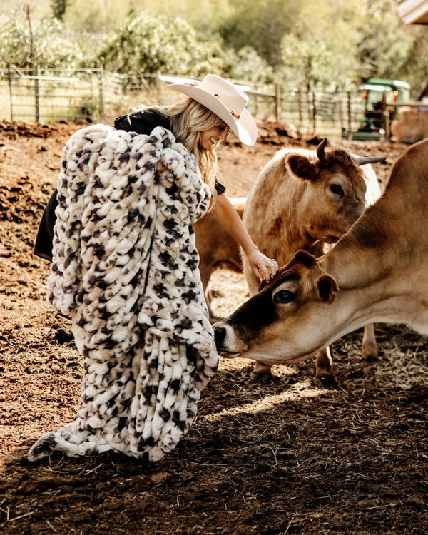 A woman in a cowboy hat and black top is wrapped in a chunky black-and-white pig-print faux fur blanket while standing in a dirt paddock with two cows.
