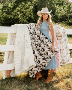 A woman wearing a cowboy hat and boots stands outdoors beside a white fence, holding a chunky black-and-white faux fur throw blanket. Three plush textured throws in cream, dusty pink, and taupe are draped over the fence behind her.