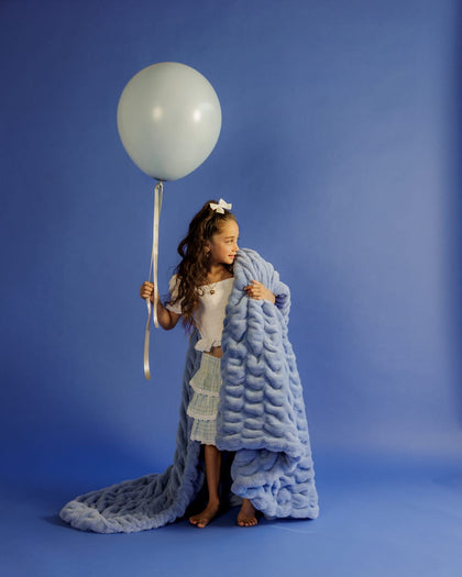 Young girl holding a large white balloon wrapped in a blue textured blanket against a blue background
