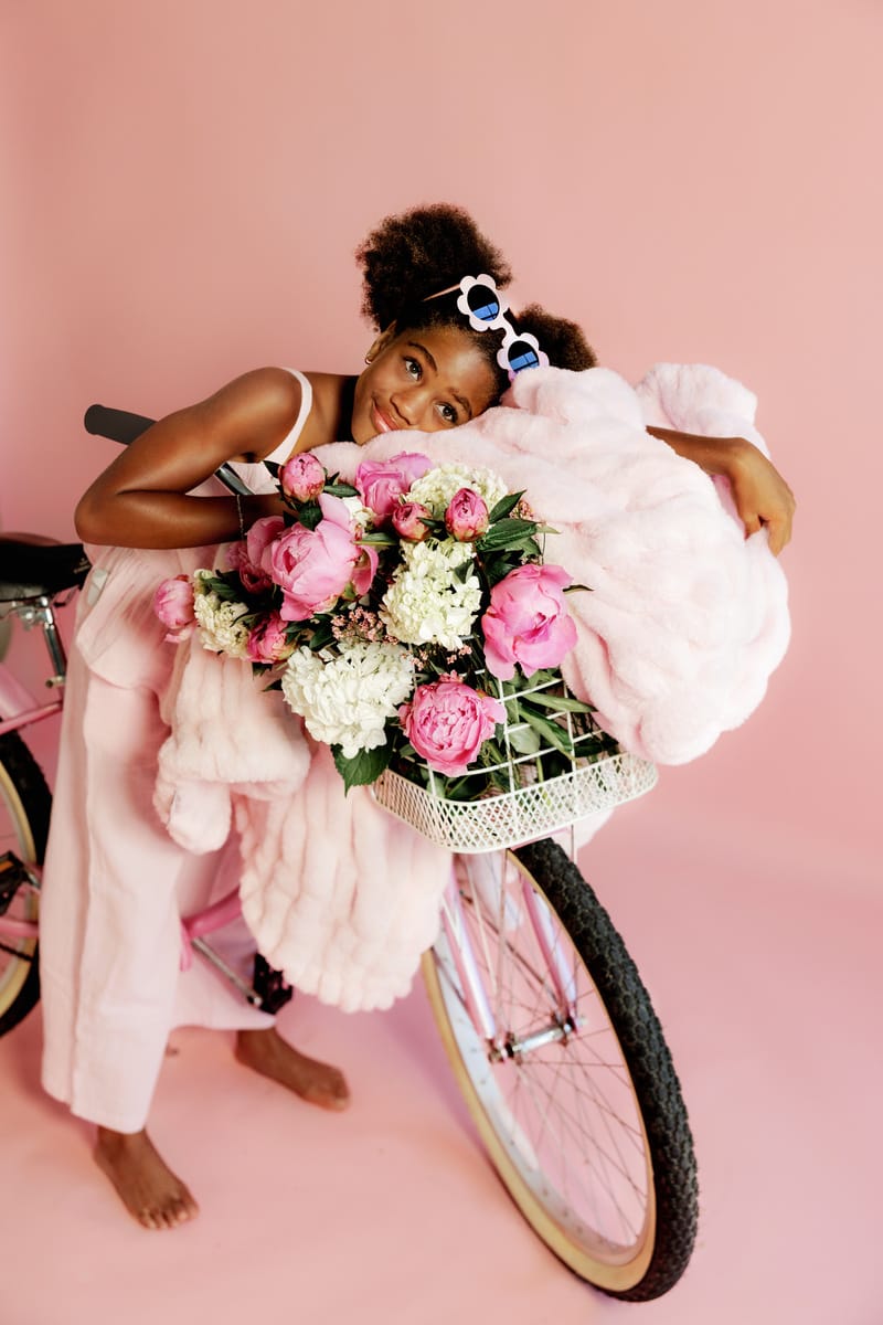 Young girl with a bicycle and flower basket with a light pink blanket on a pink background
