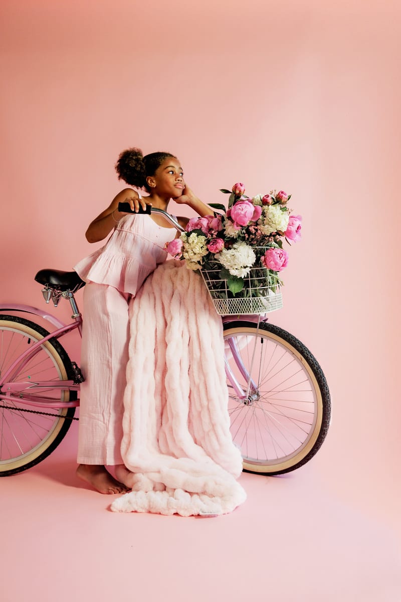 Young girl with a bicycle and flower basket with a light pink blanket on a pink background