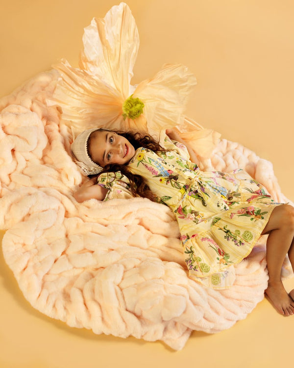 Young girl lying on a large, fluffy pastel orange blanket with a yellow dress against a beige background