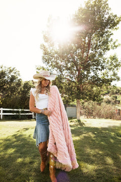 A woman outdoors in a sunny grassy yard wearing a white cowboy hat, denim skirt, white top, and brown boots while holding a chunky pink throw blanket.
