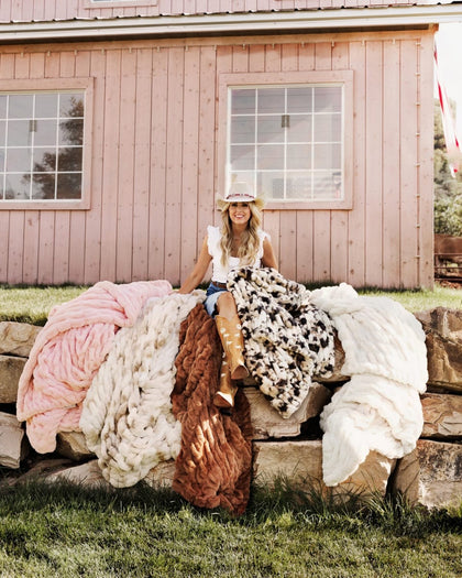 A woman wearing a cowboy hat and boots sits on a stone wall outdoors, surrounded by several throw blankets in pink, cream, brown, and black-and-white pattern. A pink wooden barn is in the background.