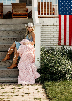 A woman wearing a cowboy hat and tall brown boots sits on outdoor concrete steps beside a white porch post, wrapped in a chunky pink blanket. 