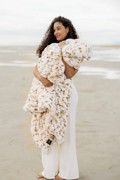 Woman holding a large, white Hugs Spotted Fawn textured blanket on a beach.
