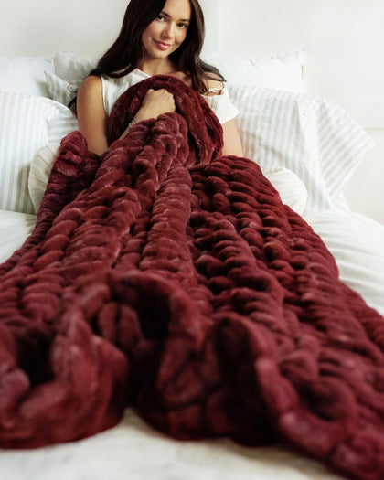 A woman sitting on a bed wrapped in a chunky, Hugs Wine Blanket. The blanket is made of a soft, plush faux fur or chenille material with a thick braided texture.