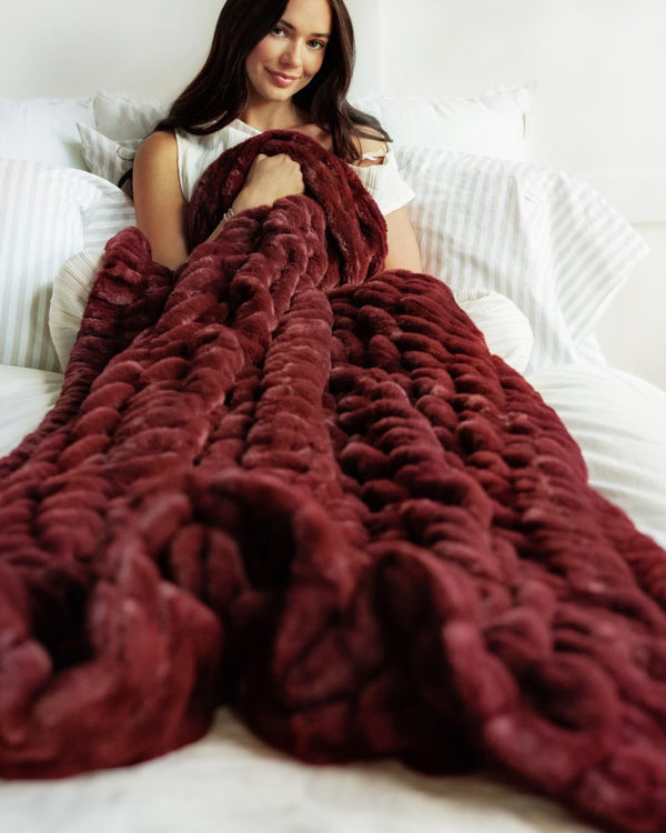 A woman sitting on a bed wrapped in a chunky, Hugs Wine Blanket. The blanket is made of a soft, plush faux fur or chenille material with a thick braided texture.