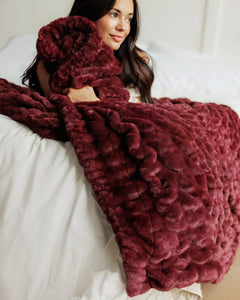 A woman lying on a bed wrapped in a chunky, Hugs Wine Blanket. The blanket has a highly textured, ruffled surface and is draped over white bedding.
