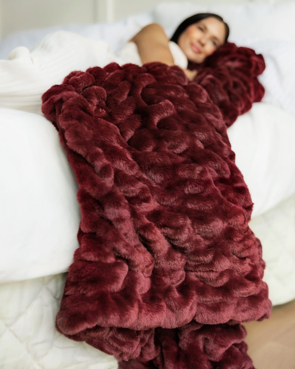 A person lying on a bed under a chunky, plush Hugs Wine Blanket. The blanket has a highly textured, wavy surface and is spread across white bedding.