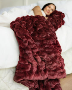 A person lying on a bed under a chunky, plush Hugs Wine Blanket. The blanket has a highly textured, wavy surface and is spread across white bedding.