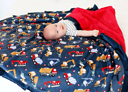 A baby lying on a Construction Zone Blanket featuring construction equipment on a navy background with a red plush minky backing.