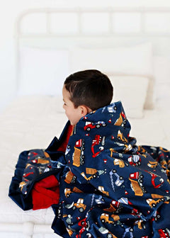 A child wrapped in a Construction Zone Blanket featuring construction vehicles on a navy blue background with a red plush backing.