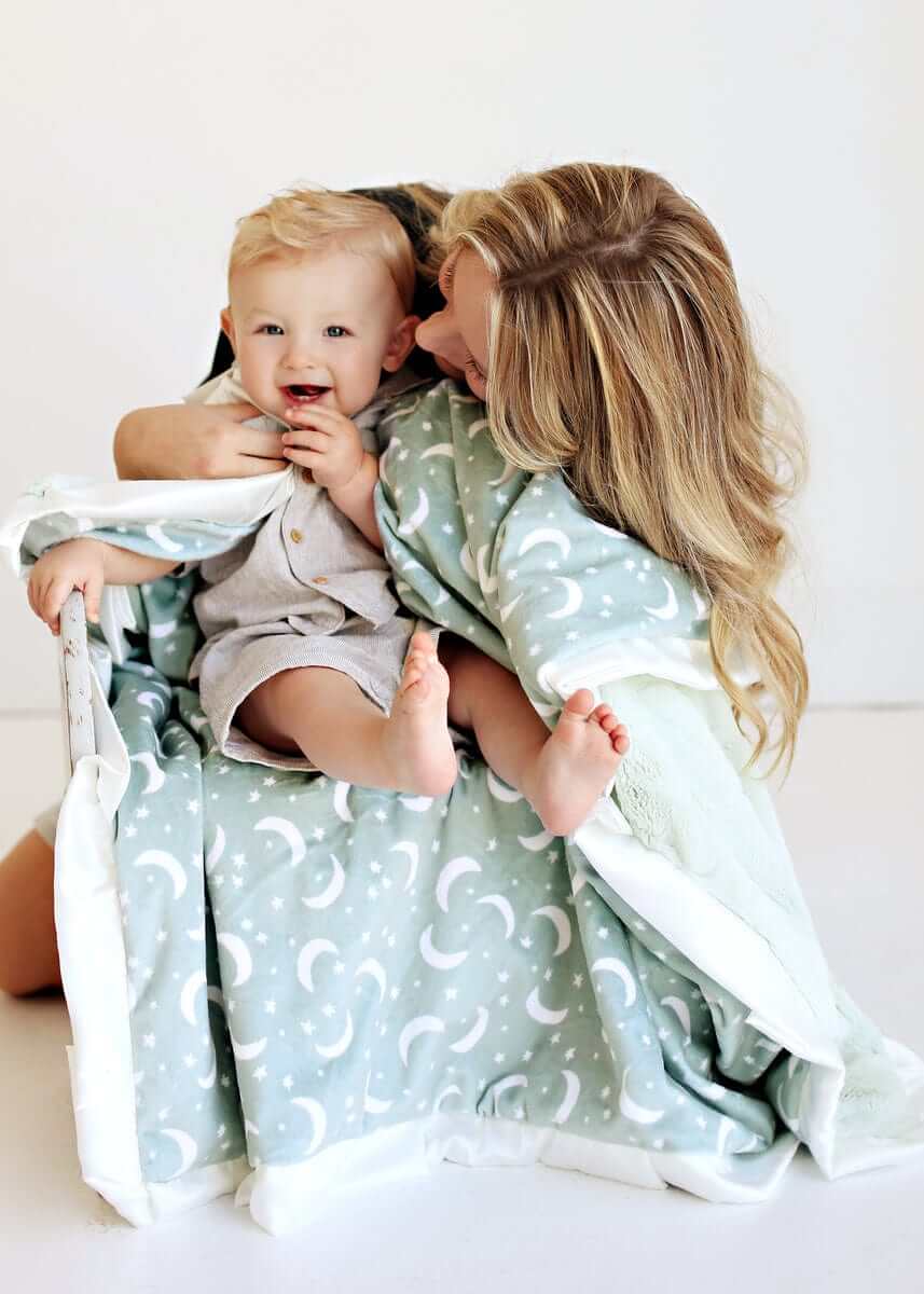 Mom snuggles with baby under the Twilight Sky Blanket featuring stars and crescent moons on sage green.