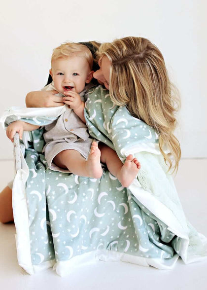 Mom snuggles with baby under the Twilight Sky Blanket featuring stars and crescent moons on sage green.