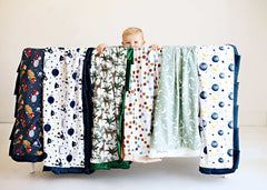Child peeking over a variety of Baby Blankets displayed, showcasing unique designs and minky fabric comfort.