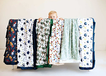 Child peeking over a variety of Baby Blankets displayed, showcasing unique designs and minky fabric comfort.