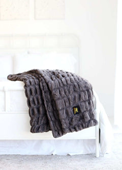 Hugs Charcoal Blanket draped over a bed, showcasing its luxurious faux fur texture and elegant charcoal gray color.
