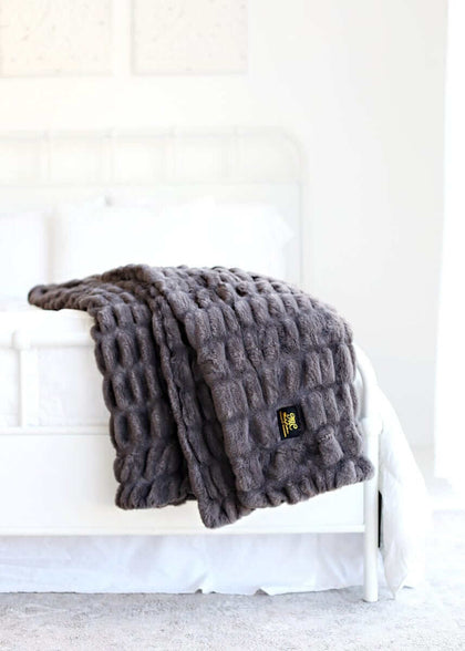 Hugs Charcoal Blanket draped over a bed, showcasing its luxurious faux fur texture and elegant charcoal gray color.