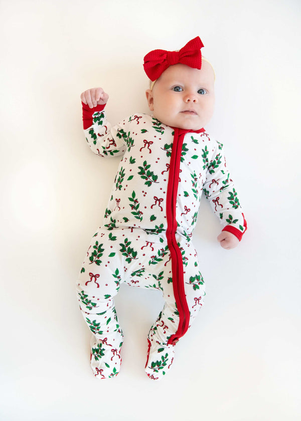 Baby wearing white holiday pajamas with holly print, red zipper, and red bow headband on a white background.