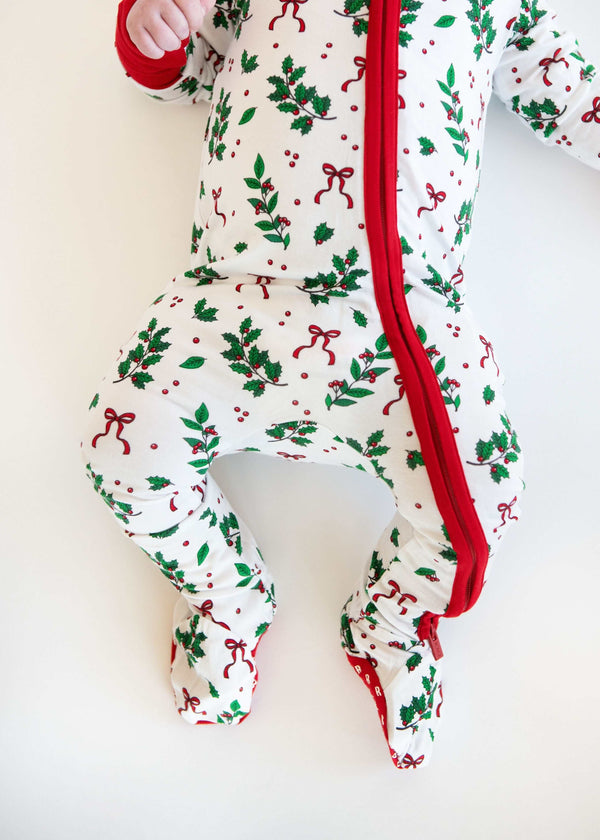 Baby wearing white holiday pajamas with holly print, red zipper, and red bow headband on a white background.