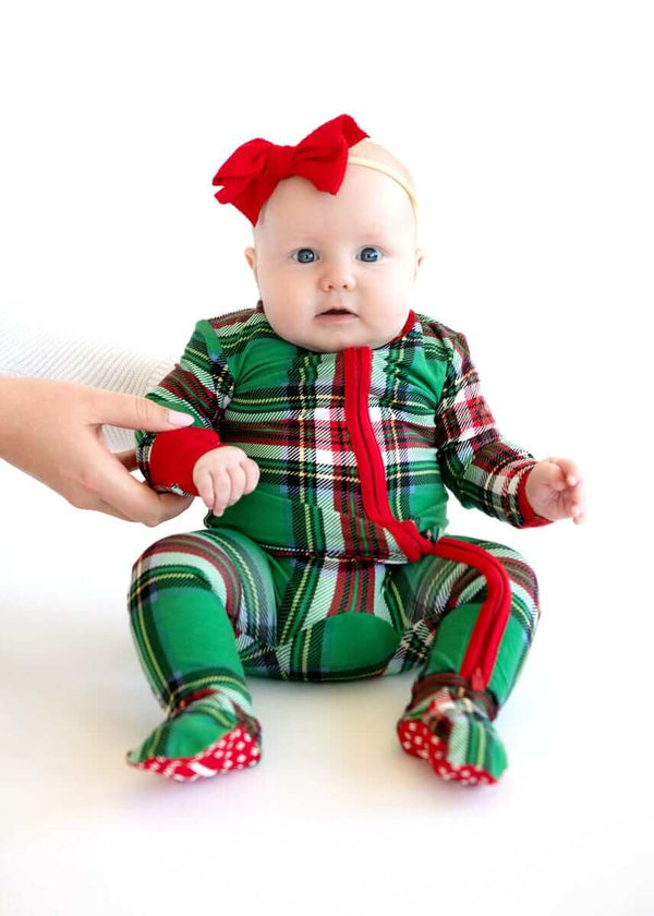 Baby wearing Minky Couture green plaid holiday pajamas with red zipper and bow headband, sitting upright on a white background.