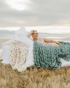 Woman sitting on a grassy field with three different textured blankets draped over her.