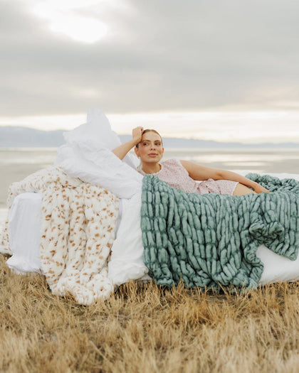 Woman sitting on a grassy field with three different textured blankets draped over her.