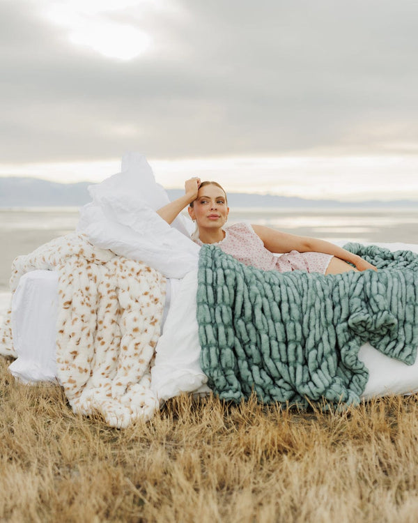 Woman sitting on a grassy field with three different textured blankets draped over her.