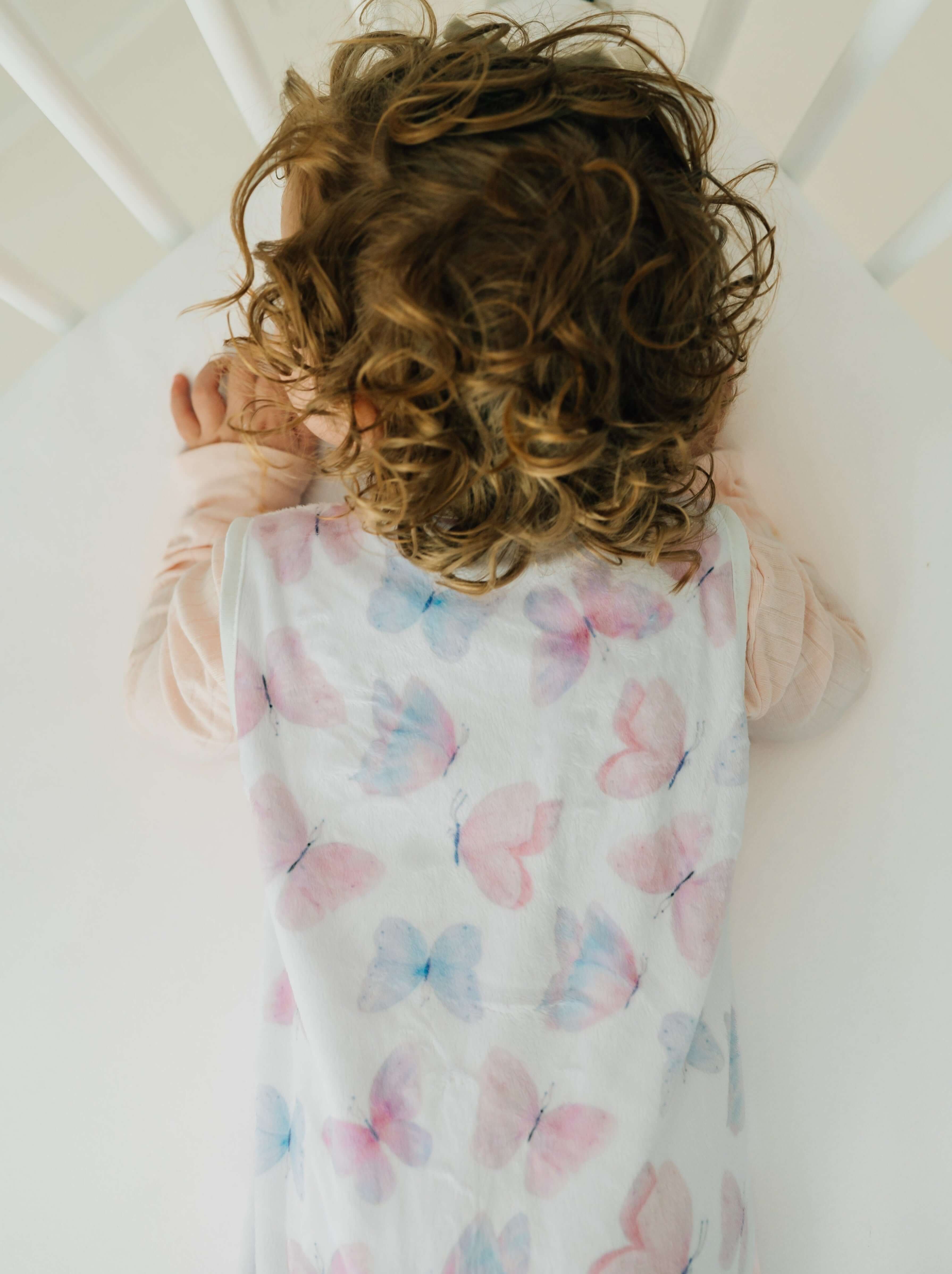 Butterfly Wearable Minky Blanket draped on a sleeping infant with curly hair in a crib.