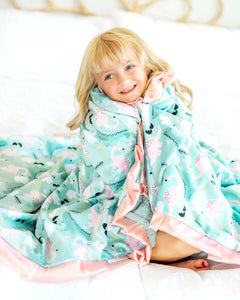 Child snuggled in Ms. Dino Blanket with blue and pink dinosaur pattern, showcasing comfort and fun design.