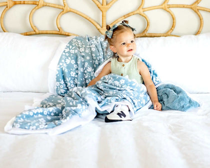 A child wrapped in a Dainty Floral Blue Blanket with small white flowers, sitting on a bed.