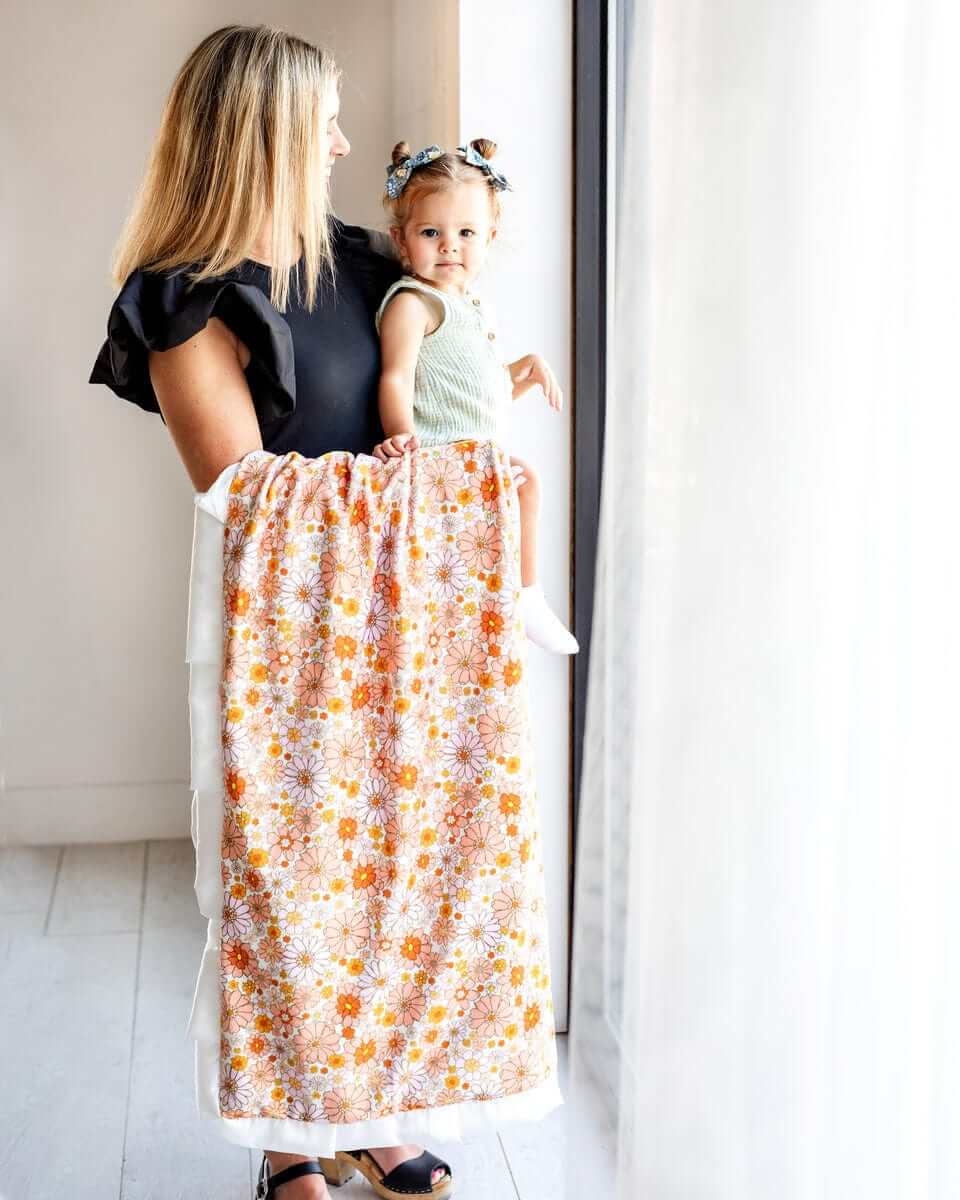 Mom holding her child while draping a Boho Floral Blanket with vibrant flowers by the window.