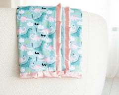 Ms. Dino Blanket featuring blue and pink dinosaur patterns on a light blue backdrop with a pink satin ruffle.