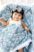 Baby wrapped in Dainty Floral Blue Blanket with small white flowers, smiling and wearing a gray bow headband.