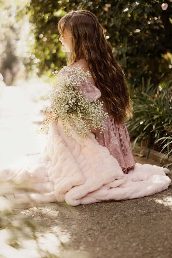 A girl with long hair sitting outdoors wrapped in a Hugs Hugs Petal Pink blanket, holding fresh flowers.