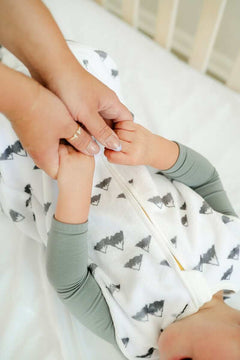 A parent gently holding a baby's hands while they wear the Mountains Wearable Minky Blanket in a crib.