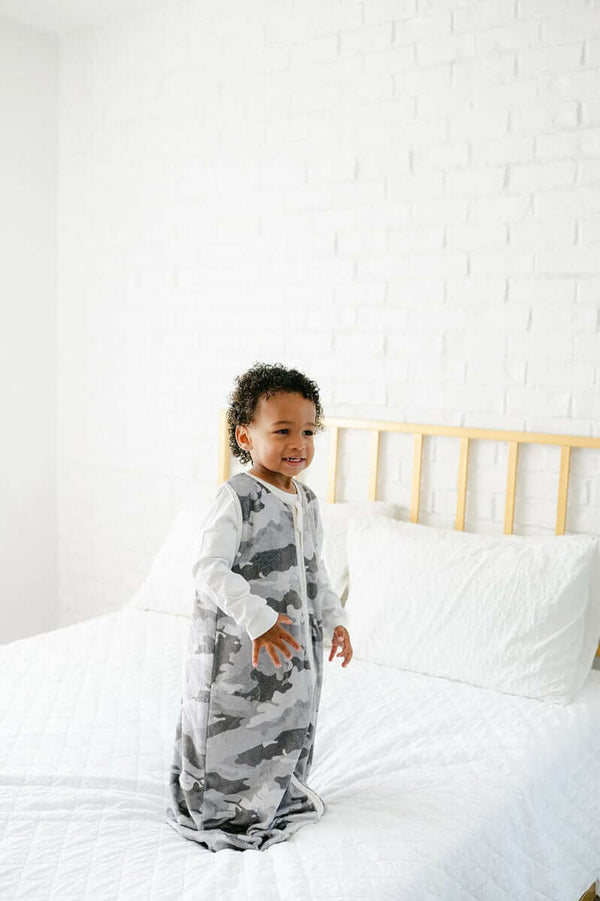 Toddler wearing a Camo Wearable Minky Blanket, smiling and playing on a bed with white bedding.
