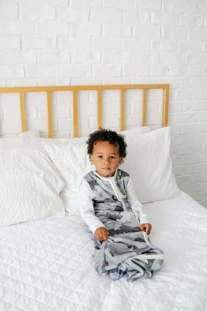 Child comfortably wearing a Camo Wearable Minky Blanket, featuring a gray camo pattern and a gold zipper, sitting on a bed.